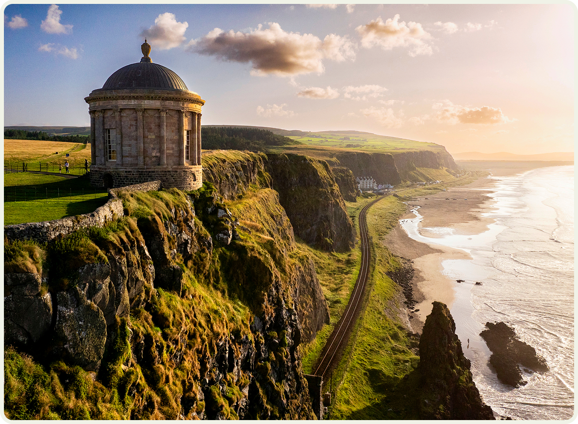 Coastal cliffs at sunset with a small stone temple overlooking the ocean and winding railway below.